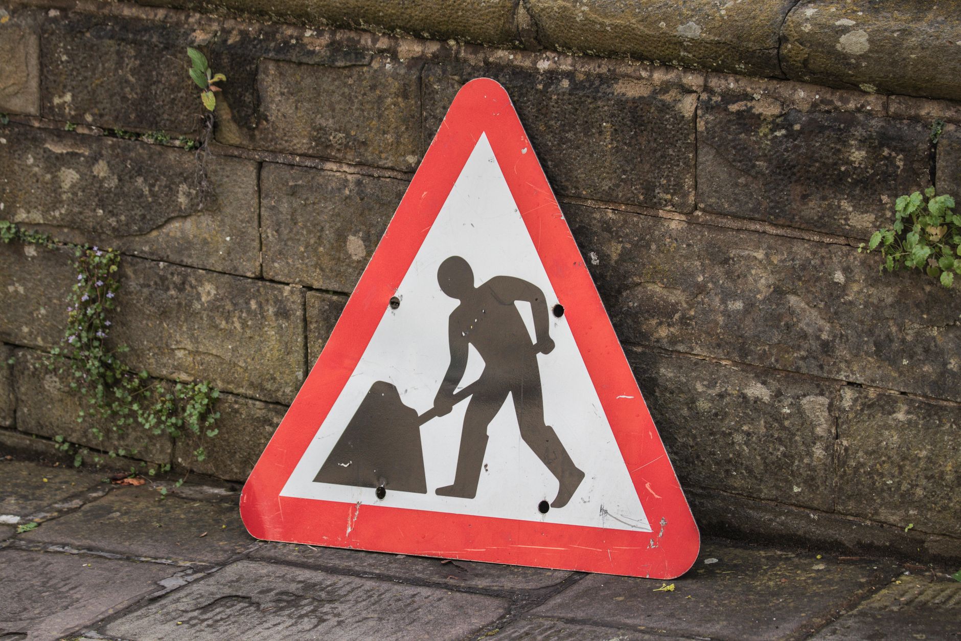 Triangular road work sign placed on a city sidewalk beside a stone wall.