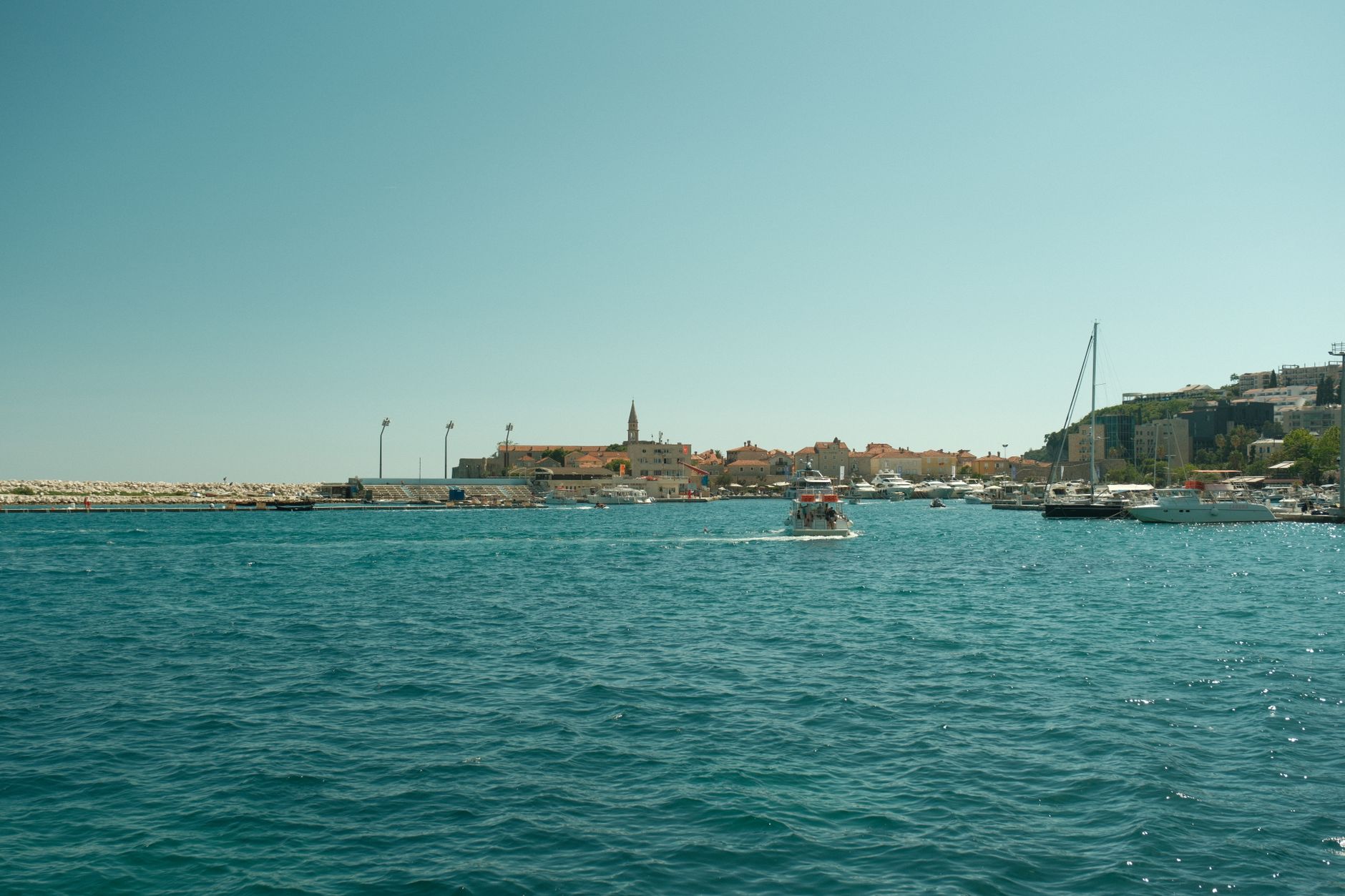 A vibrant summer view of Budva's harbor with boats on the Adriatic Sea under a clear sky.