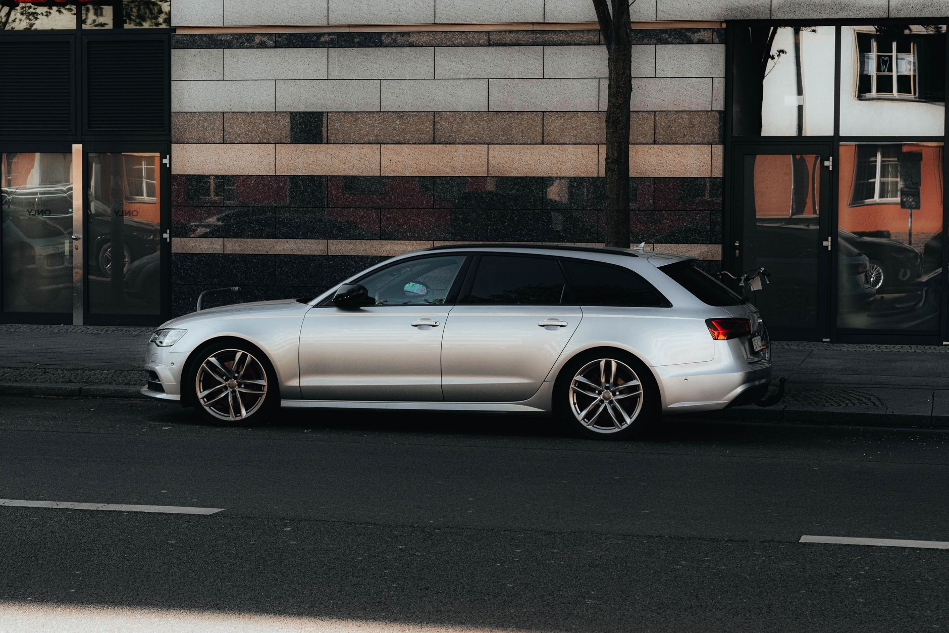 A silver luxury car parked on a Berlin street with modern architecture reflecting in city windows.