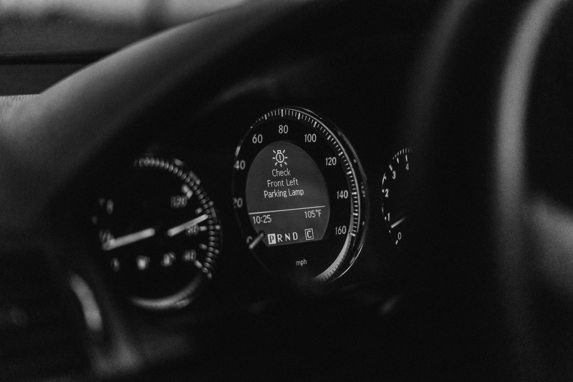 Close-up of a car dashboard in black and white with a parking lamp warning light.