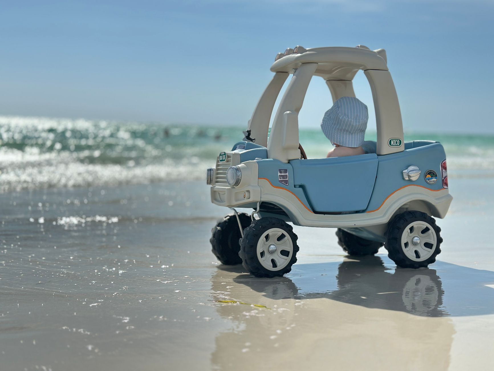 A toddler in a toy car enjoys a sunny day at the beach, capturing the essence of fun and adventure.