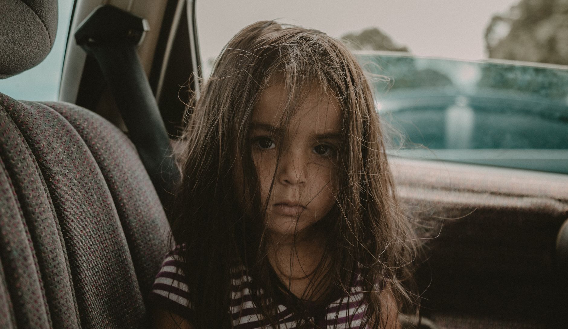 A young girl with long, messy hair sitting in a car seat, looking directly at the camera.