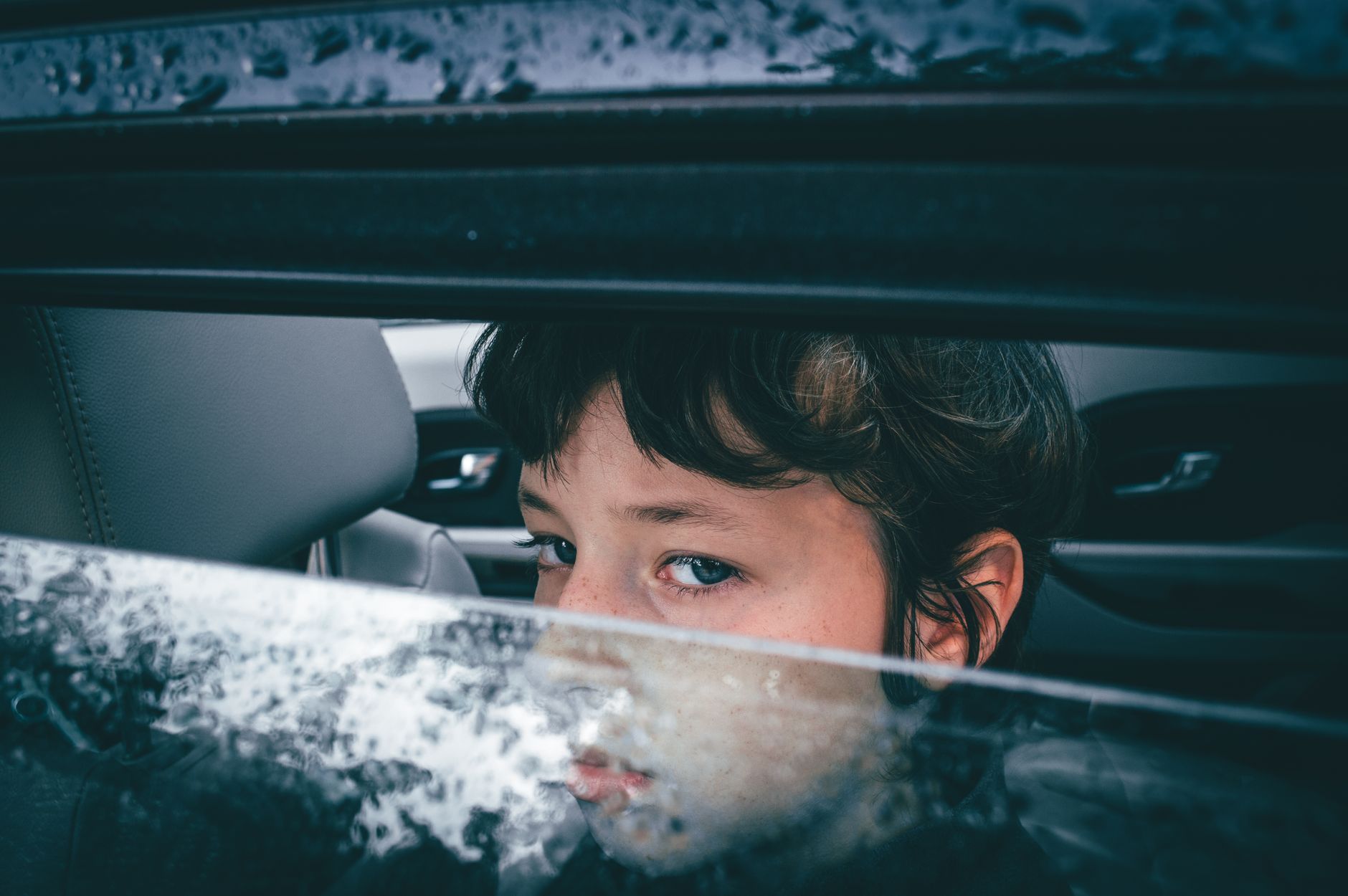 Close-up of a child looking thoughtfully through a rain-covered car window, evoking a sense of introspection.