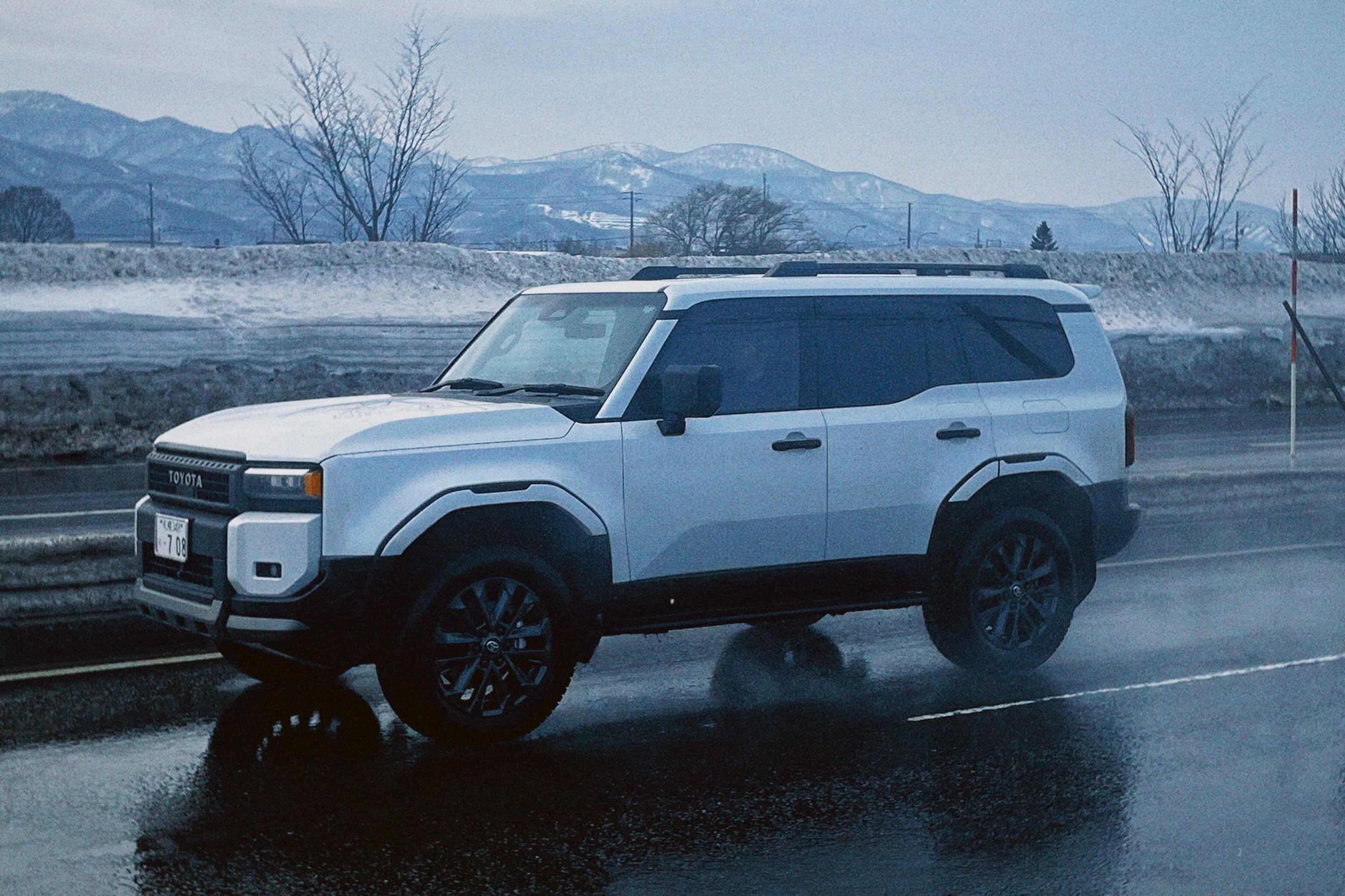White SUV driving on a wet road against a snowy, mountainous backdrop in Sapporo, Japan.