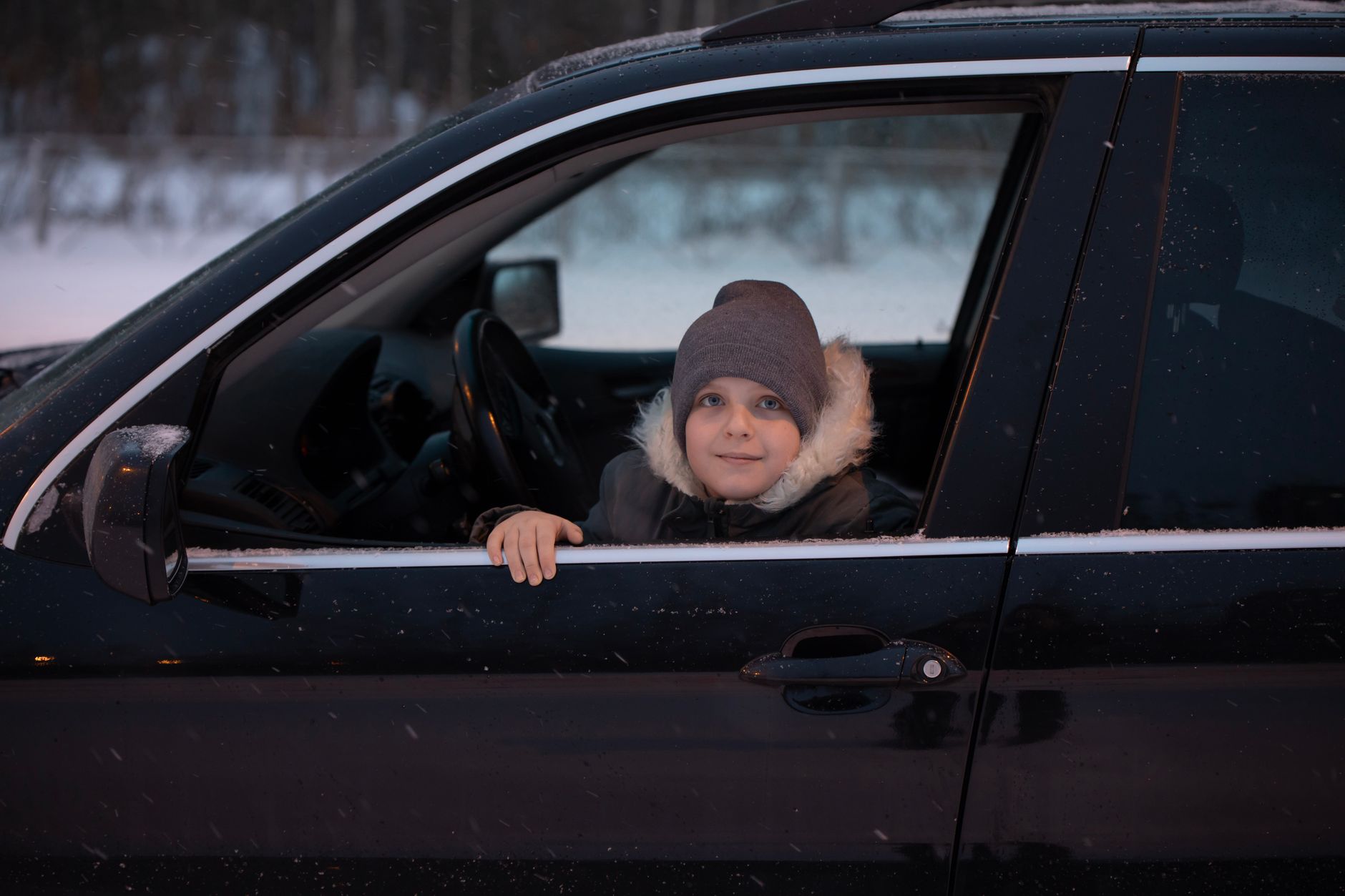 A young child in warm winter attire sits in a car, enjoying a snowy day outdoors.