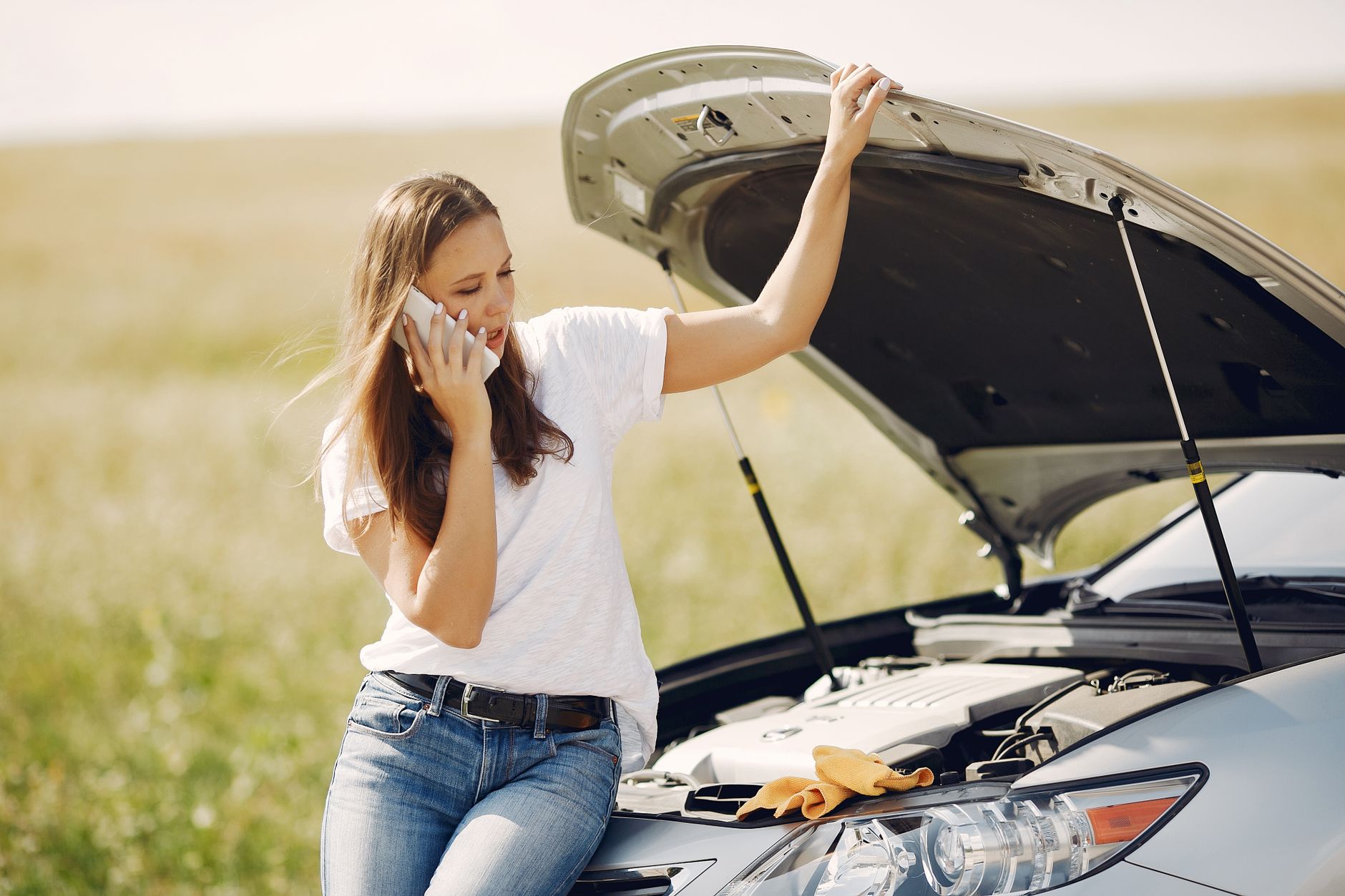 Worried young female driver in white t shirt and jeans speaking on phone while leaning on broken car with open hood asking for help with rep