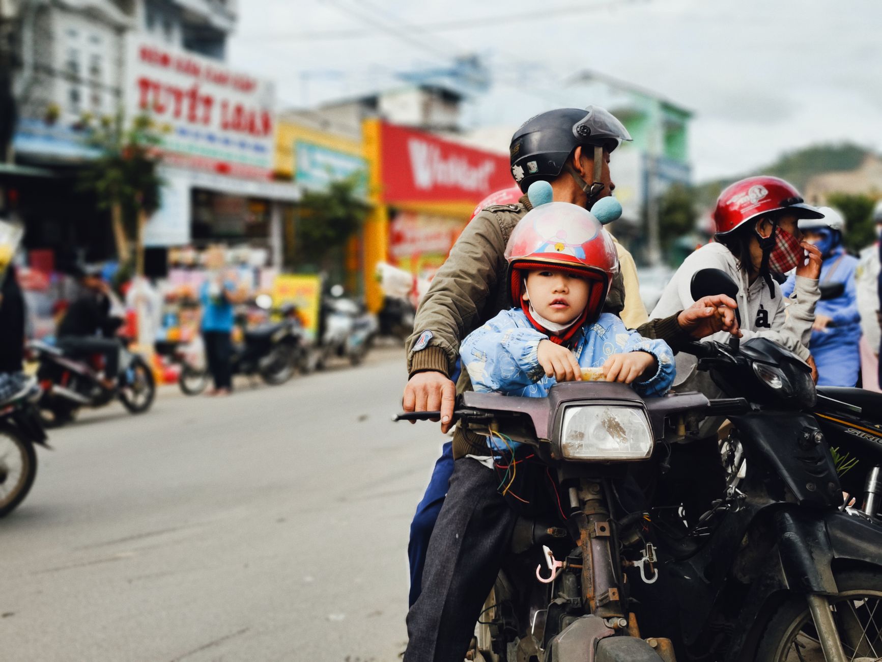 Young child on motorcycle with adult in vibrant street setting, helmets on.