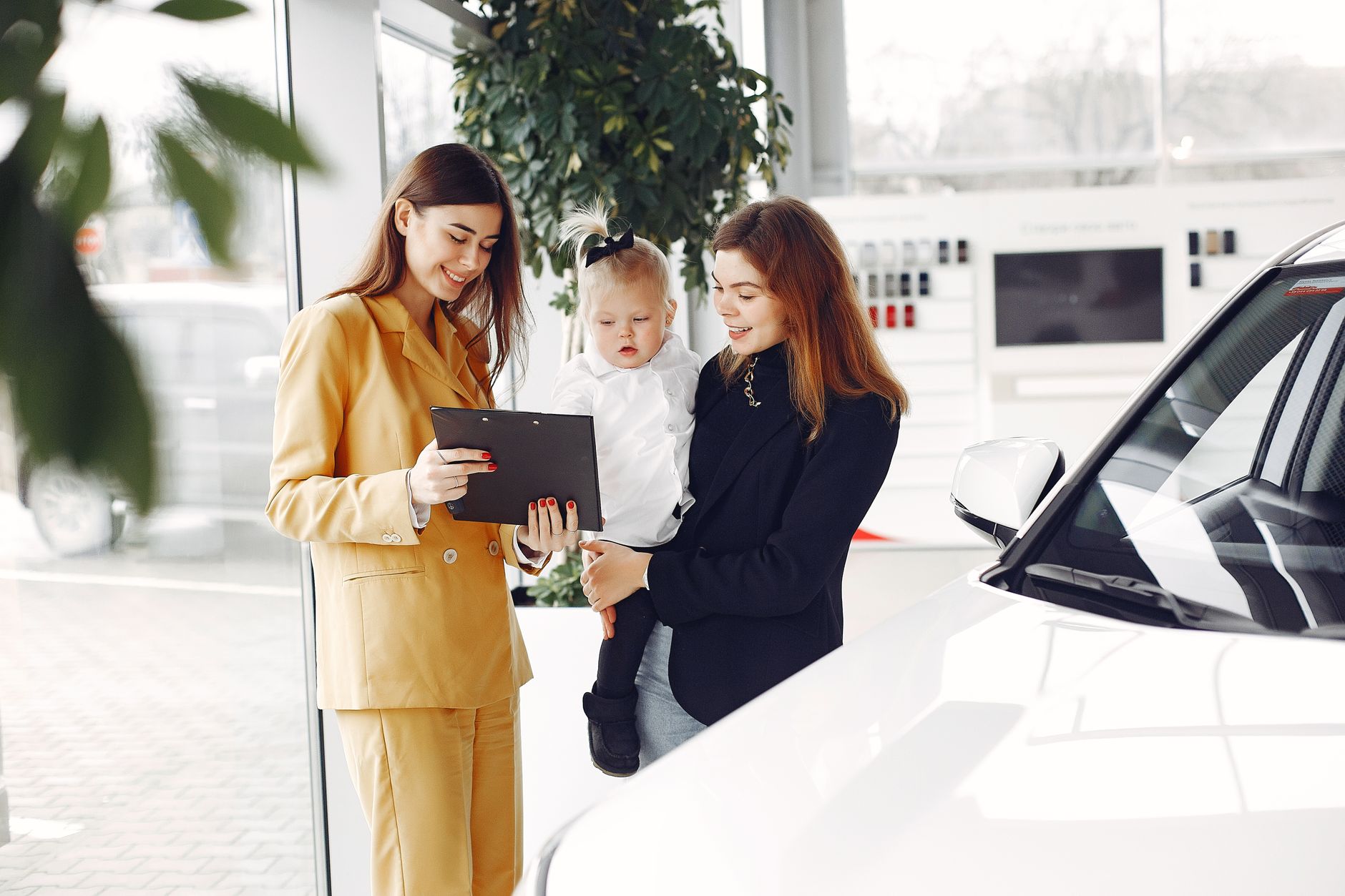Positive young female with cute little daughter on hands discussing car characteristics with professional dealer in stylish beige suit while
