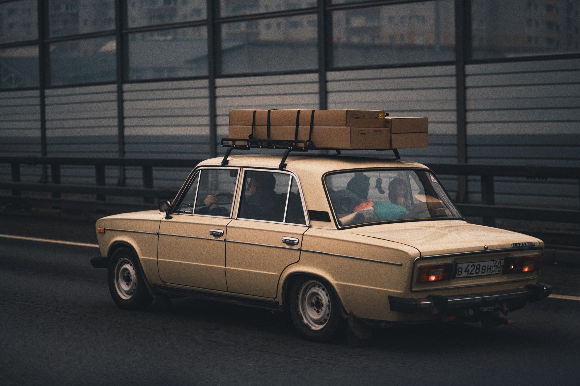 Classic beige sedan carrying luggage, driving on highway with city in background.
