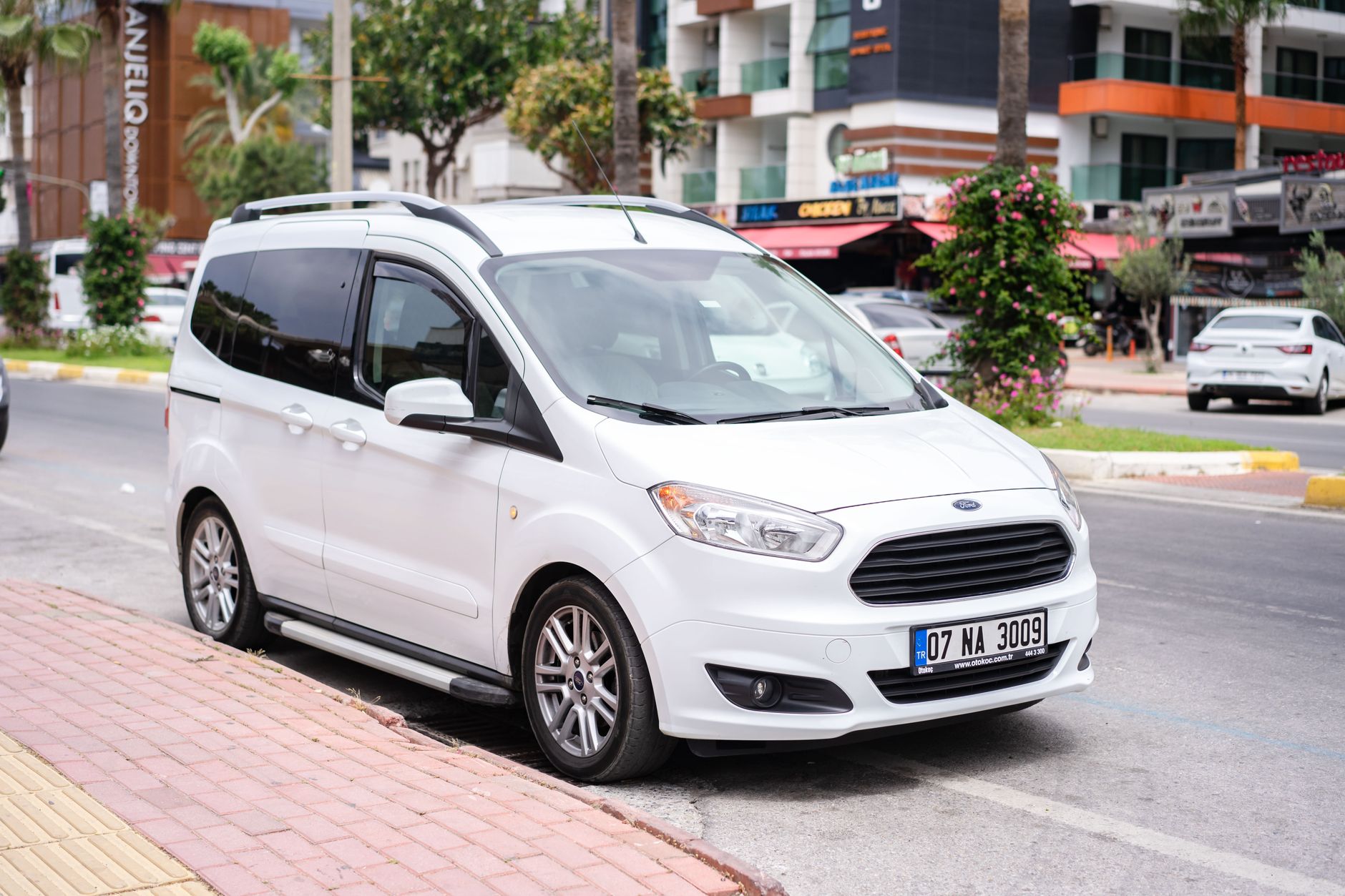 White minivan parked on a city street with modern buildings in background, illustrating urban transportation.