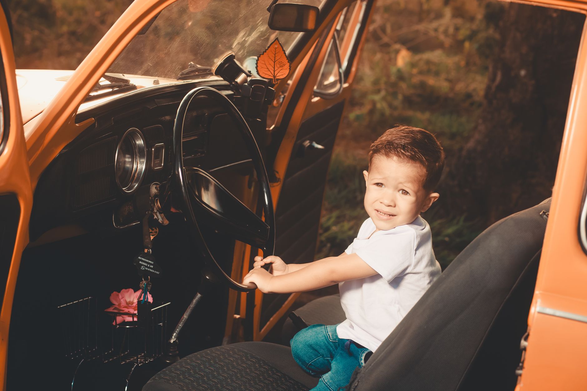Cute child sitting in an orange vintage car, pretending to drive while looking at the camera.