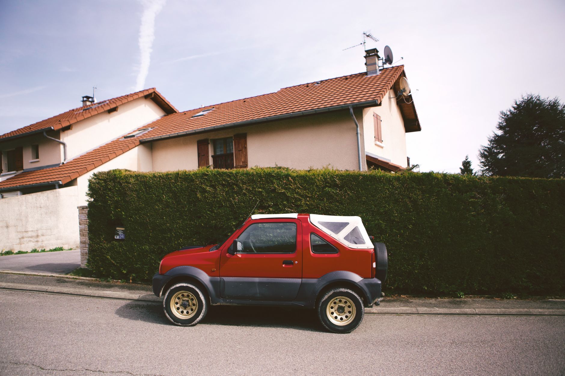 A small red SUV parked on a street in Auvergne-Rhône-Alpes, France, beside a residential house with a hedge.