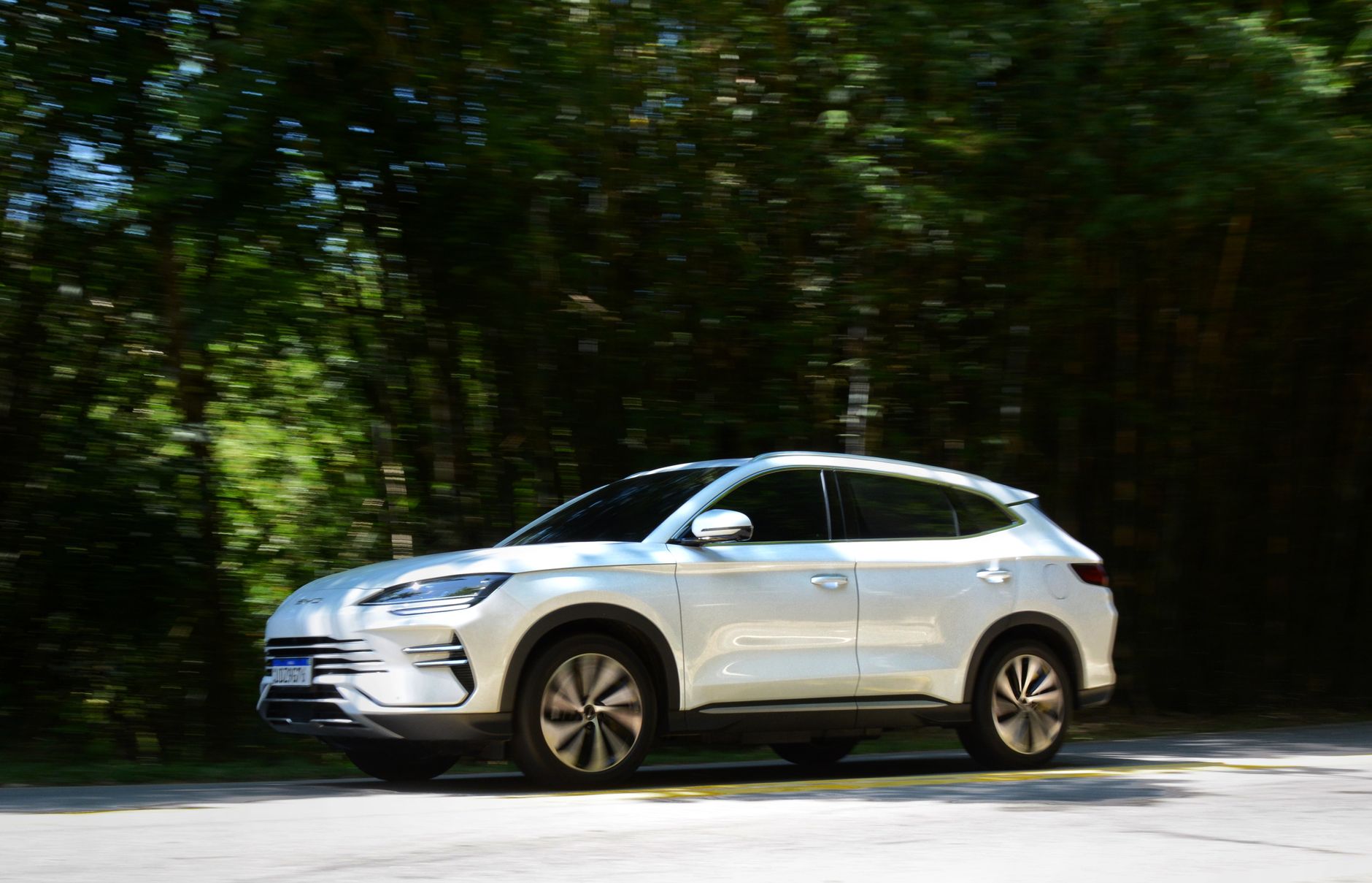 A modern white SUV driving through a lush, green forest road in Monteiro Lobato, Brazil. Daytime scene.