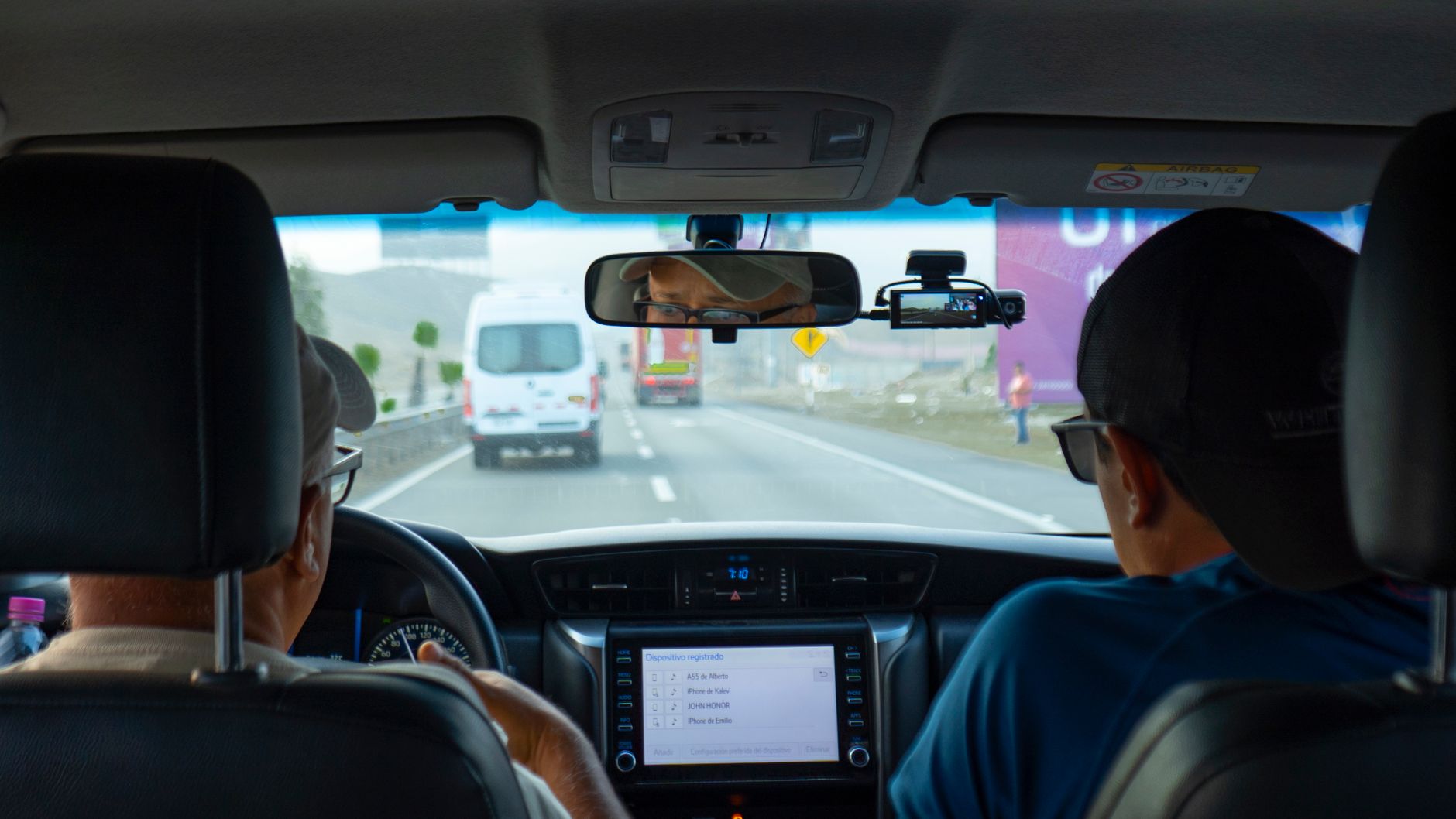 Interior of a car with two people driving on a highway, focusing on dashboard and rear view mirror.