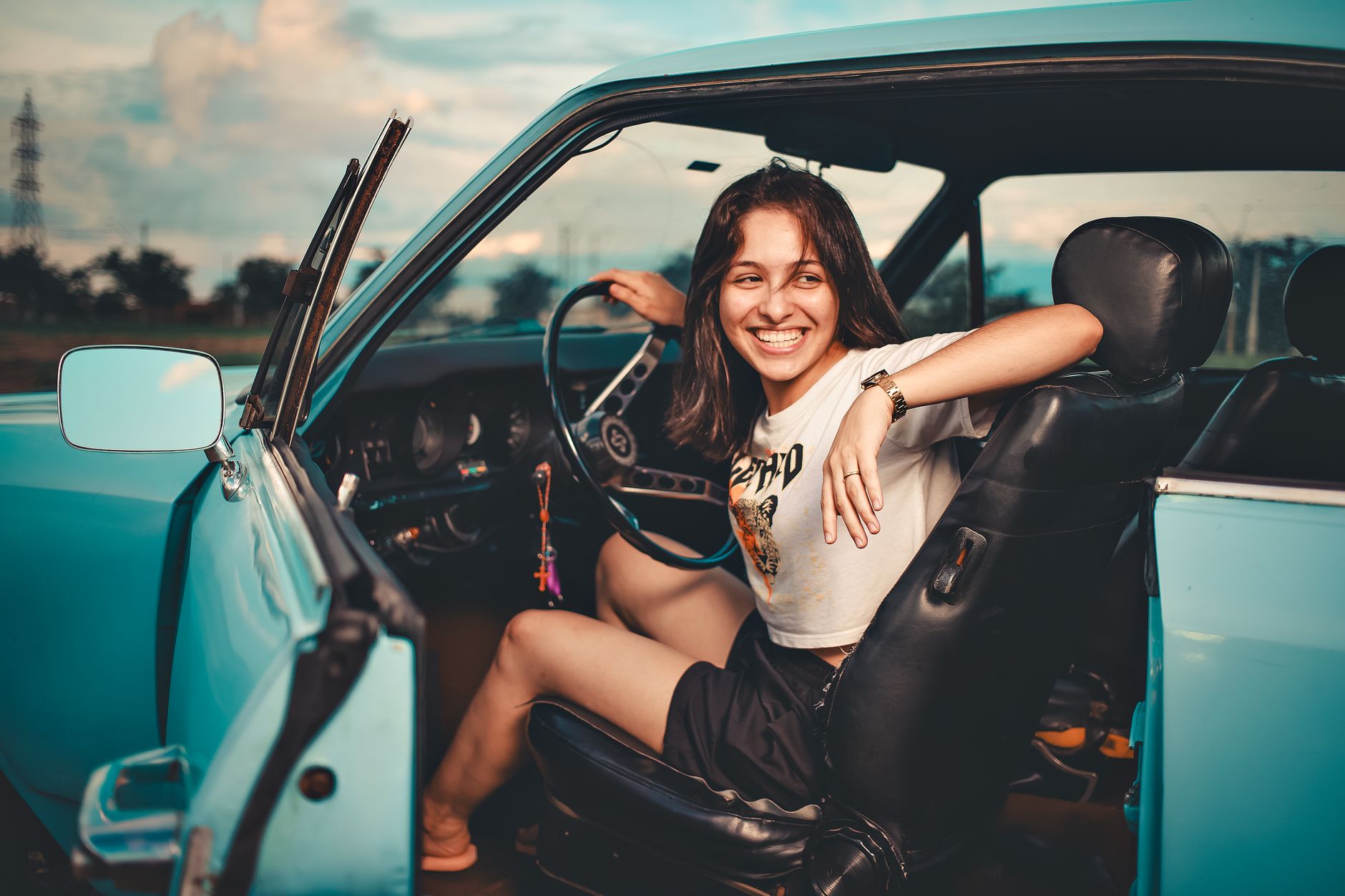A cheerful young woman sitting in a blue vintage convertible car outdoors during the day.
