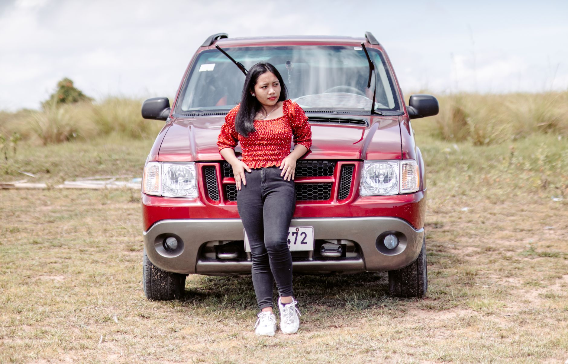 A young woman in fashionable attire poses confidently against a red SUV in a grassy field, conveying adventure.