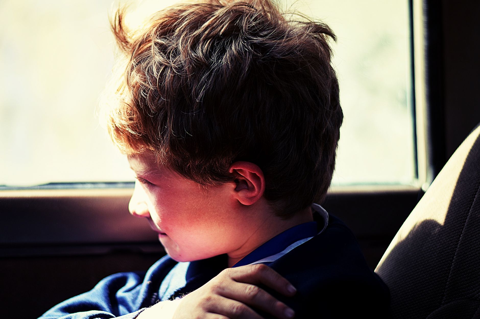 A young boy sits in a car, gazing out the window with a thoughtful expression in sunlight.