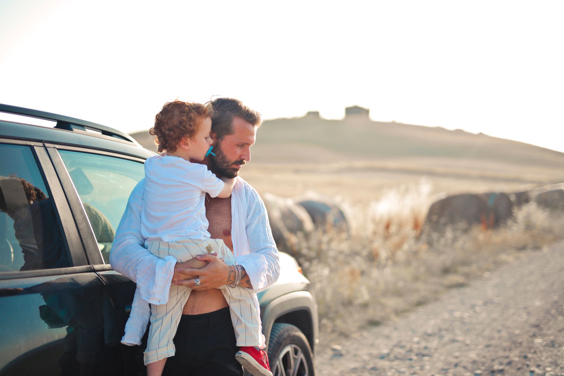 A father lovingly holds his son by a car in a peaceful countryside setting.